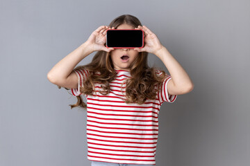 Little girl in T-shirt covering eyes with cellphone, posing with mouth wide open from shock surprise, unknown child hiding face with mobile phone. Indoor studio shot isolated on gray background.