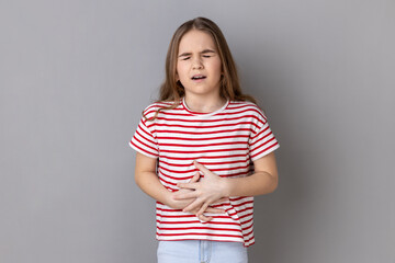 Portrait of little girl wearing striped T-shirt clutching belly, feeling discomfort or pain in...