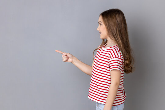 Side view portrait of little girl wearing striped T-shirt standing pointing aside presenting copy space for advertisement or promotional text. Indoor studio shot isolated on gray background.