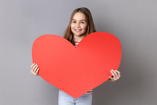Portrait of cute little girl wearing striped T-shirt holding big red heart and smiling to camera, child expressing love and fondness for parents. Indoor studio shot isolated on gray background.