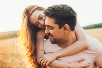 Fototapeta premium Portrait of smiling handsome man giving his ginger girlfriend a piggy ride back in wheat field. Beautiful portrait. Leisure activities. Summer activities