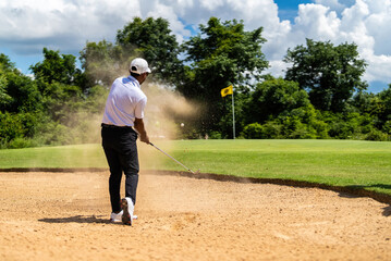 Asian man golfing on the course. He hits a golf ball on a sand field.
