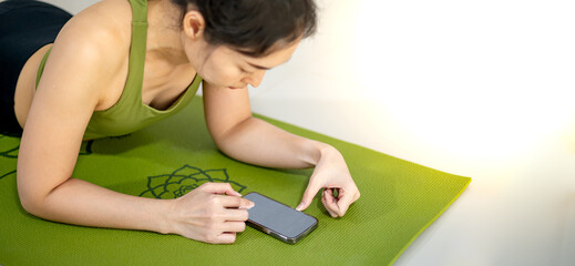 Women play mobile and sitting on the green yoga mat while resting practice in the home.
