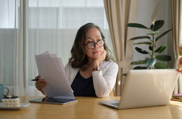 Thoughtful senior mature woman watching business training, online webinar on laptop computer while...