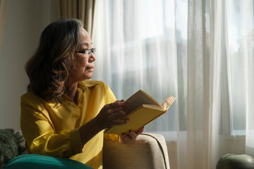 Happy elderly woman taking a break from reading while looking through the window. Retirement concept.