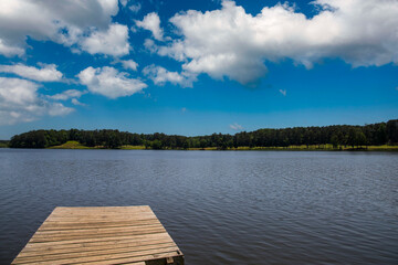 Fototapeta na wymiar a long square brown wooden dock over the rippling waters of Lake Acworth surrounded by lush green trees and grass with a gorgeous blue sky and clouds at Cauble Park in Acworth Georgia USA 