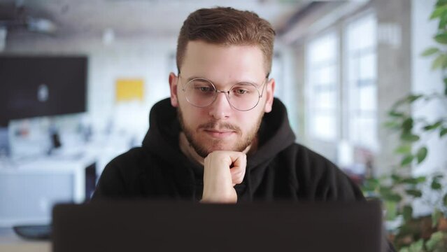 Thoughtful professional programmer sitting and pondering in front of laptop monitor in office. Skilled attentive male worker coding updates to multiplayer game.