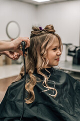 Fototapeta premium Cute child getting hair done. Hairdresser with curling iron. Generation of fashion.
