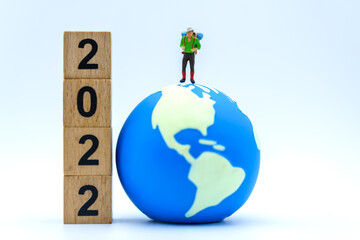 2022 New Year and Travel Concept. Closeup of traveler miniature figures with backpack standing on mini world ball  with stack of wooden number block on white background.