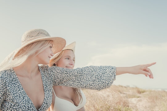 Two Teenage Girls Enjoy A Sunny Day On The Mediterranean Coast During Their Summer Vacation. They Look Far Away Pointing Something At The Horizon. Friendship Concept.