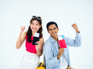 Ready to travel, happy holiday. Summer vacation concept. Smiling Asian couple holding raised fist, showing passport with success, young woman and man sit on suitcase isolated on white background.