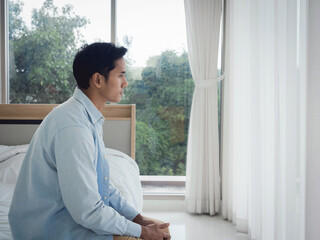 Thoughtful Asian man in denim shirt sitting on the bed feeling lonely, looking away with sadness and thinking in the bedroom at home near the glass window in the morning.