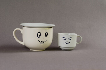 Two ceramic cups against a gray background. A large cup with a smiling woman's face painted on it. A small cup with a man's face. Heterosexual couple. Flirt, love, relationship concept.
