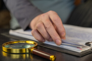 A man's hand is leafing through documents. Round magnifying glass with gold-colored frame in the foreground. A clipboard for papers. Inside the room. Selective focus.
