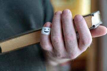 A man is reading a book while holding it in his hands. The first phalanx of his pinky finger is wrapped in white ribbon. A smiling face is painted on the bandage.