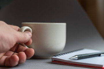 Fototapeta na wymiar Close-up of a man's hand holding a beige ceramic cup with a handle. Open spring-loaded notebook and pen lying side by side. Side view. Selective focus.