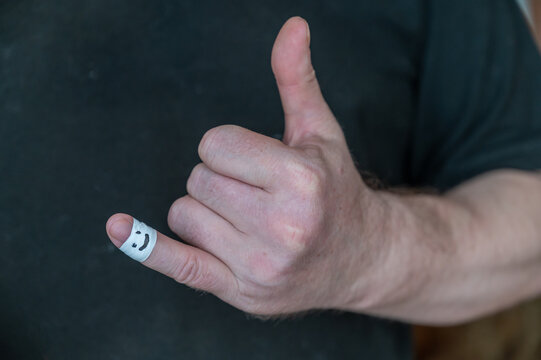 The man shows his hand a thumbs-up gesture. The first phalanx of his little finger is wrapped in white ribbon. A smiling face is painted on the bandage. Positive emotion concept. Selective Focus.