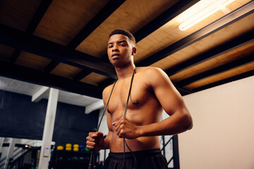 Fototapeta na wymiar African American male looking serious into the camera. Topless mixed race male exercising indoors in the gym with skipping rope. High quality photo