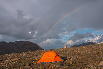 Scenic mountain landscape with vivid orange tent in sunlight and rainbow in cloudy sky. Beautiful alpine scenery with tent under rainbow in overcast. Orange tent and high rainbow in rainy weather.