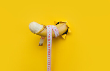 A banana with a peeled penis-shaped peel and a tape-shaped ruler is pushed through a torn hole in...
