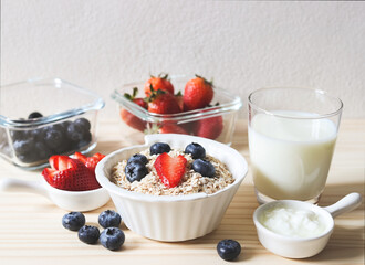 Front view of breakfast with oat or granola in white bowl, fresh blueberries, strawberries, a  glass of milk on wooden table. Healthy breakfast concept.