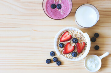  flat lay of  breakfast with oat or granola in white bowl, fresh blueberries, strawberries, a  glass of milk, smoothie blueberries  and yogurt on wooden table. Healthy breakfast concept.