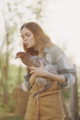 A happy young woman smiles at the camera and holds a young chicken that lays eggs for her farm in the sunlight