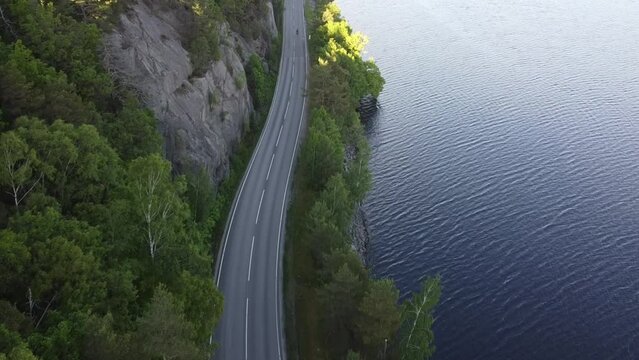 Aerial footage of a curvy road and driving motorcycle.  Drone movie taken from above in Sweden in summer. Surroundings with trees and a lake. Travel and transportation concept.