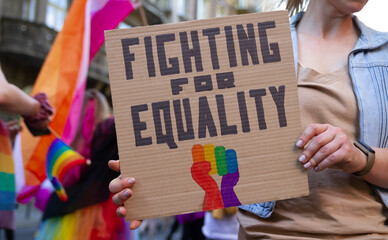 Woman holding placard sign Fighting for Equality with rainbow flag fist, during LGBT Pride Parade. Crowd of people at equality march to support and celebrate LGBT+, LGBTQ gay and lesbian community. - obrazy, fototapety, plakaty