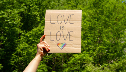 Hand holding placard sign Love is Love with rainbow flag heart, symbol of LGBT. Pride Parade,...
