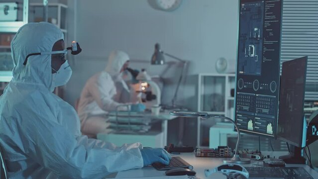 Waist up slowmo of electronics engineer in protective coveralls and respiratory mask working on computer while studying circuit board and other computer elements in lab