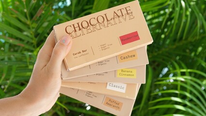 Hand holding healthy chocolate alternative carob bars with flavors. Tropical palms in the background