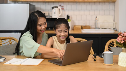 Smiling little asian girl learning internet online class and do homework with her mother in kitchen. E-learning education concept