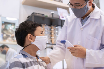 Obraz premium Masked optometrist uses a non-contact infrared thermometer gun to the boy before undergoing an eye exam, disease prevention concept