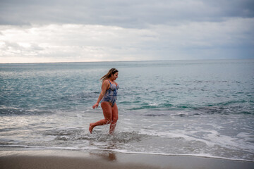 Fototapeta premium A plump woman in a bathing suit enters the water during the surf. Alone on the beach, Gray sky in the clouds, swimming in winter.