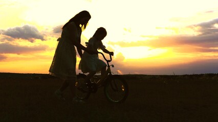 Fototapeta na wymiar young mother teaches child ride bike sunset. Little kid bike. Childhood dream concept. silhouette happy family park. mother with child girl rides bicycle pedaling sun. happy kid rides bike park.