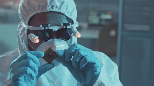 Slowmo closeup of male technician in protective coveralls, respiratory mask, gloves and special magnifying eyewear inspecting computer chip in lab