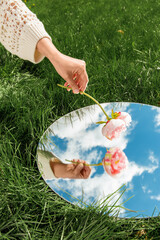 nature concept - hand with peony flower and sky reflection in round mirror on summer field