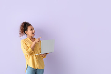 Surprised young African-American woman with laptop pointing at something on lilac background