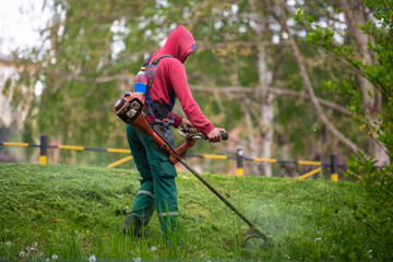 Worker mowing tall grass with electric or petrol lawn trimmer in city park or backyard. Gardening care tools and equipment. Process of lawn trimming with hand mower.