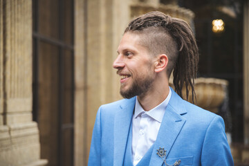 Caucasian smiling young man with dreadlocks in blue business suit, outdoor portrait.