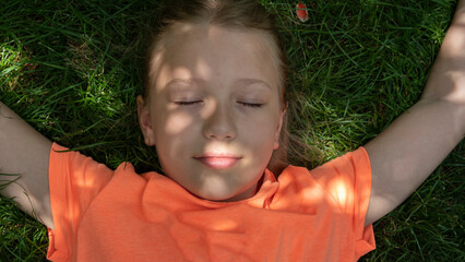 Smiling girl with closed eyes lies on bright green grass.  Happy childhood