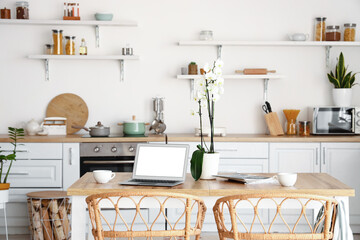 Modern laptop, cups and orchid flower on dining table in kitchen