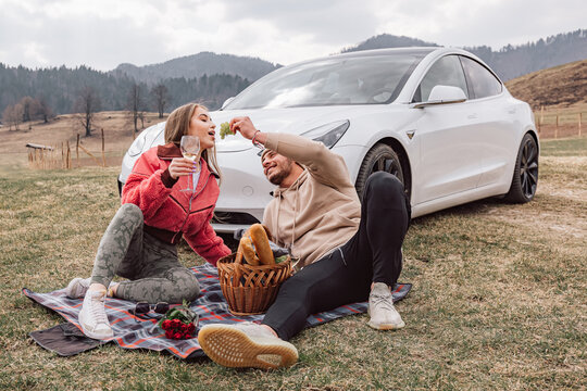 Young modern couple enjoying a picnic in nature, sitting near the parked white electric car