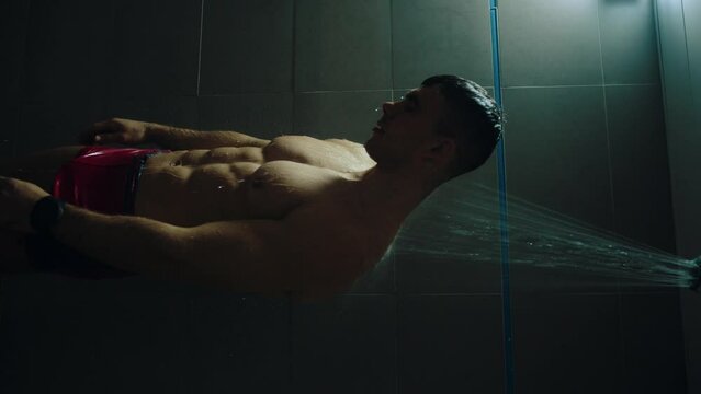 Young muscular man in red swimming trunks washes himself in the public shower of the gym or swimming pool. Vertical video