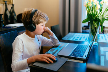 Focused caucasian teenage boy with headphones using laptop at table in room. Cute preteen boy doing...
