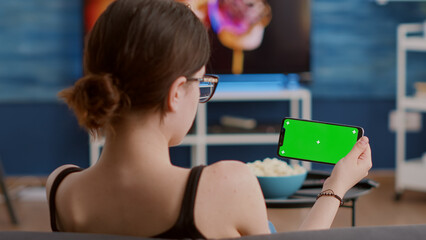 Young woman holding horizontal smartphone with green screen using streaming app watching video content in living room. Girl using touchscreen mobile phone with chroma key in group video call.