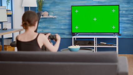 Static tripod shot of gamer girl holding wireless gamepad playing action console video game on green screen tv while sitting on sofa. Woman enjoying online gaming on chroma key display with controller © DC Studio