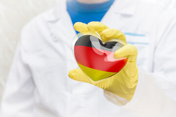 A doctor in a medical gown and protective gloves holds a heart in the colors of the German flag in front of him. The face is not visible.