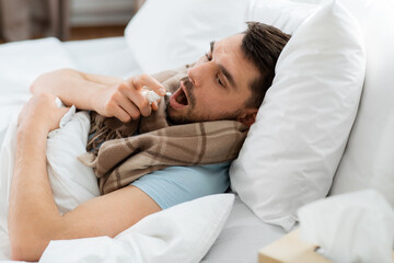 people and health problem concept - unhappy sick man spraying his throat with oral spray lying in bed at home